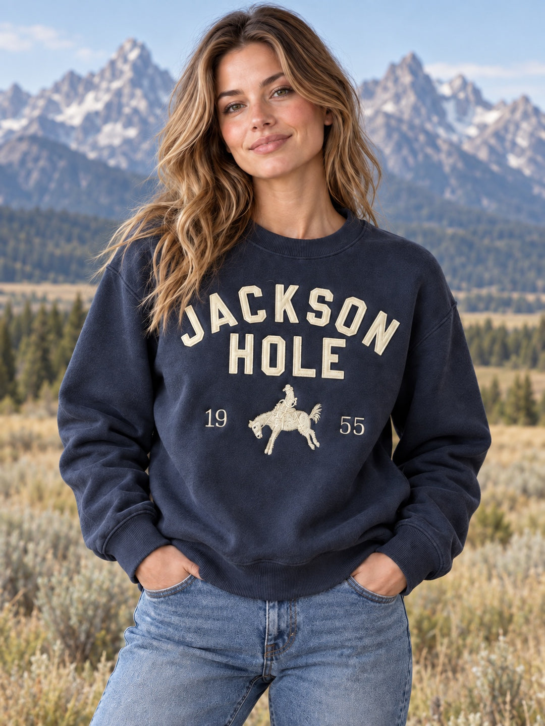 Woman wearing the navy Jackson Hole 1955 sweatshirt with cream lettering and bronco rider, Teton mountains behind